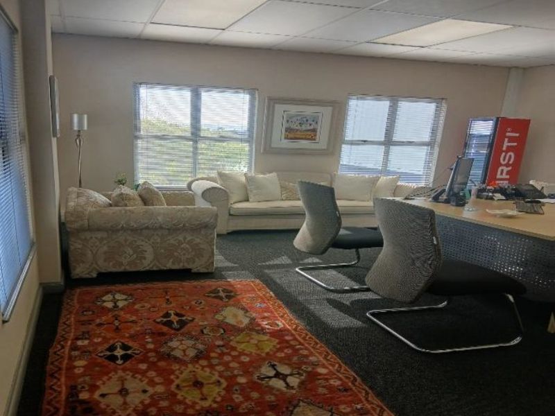A functional office space featuring carpeted flooring, a suspended ceiling with recessed lighting, and large windows with venetian blinds.
