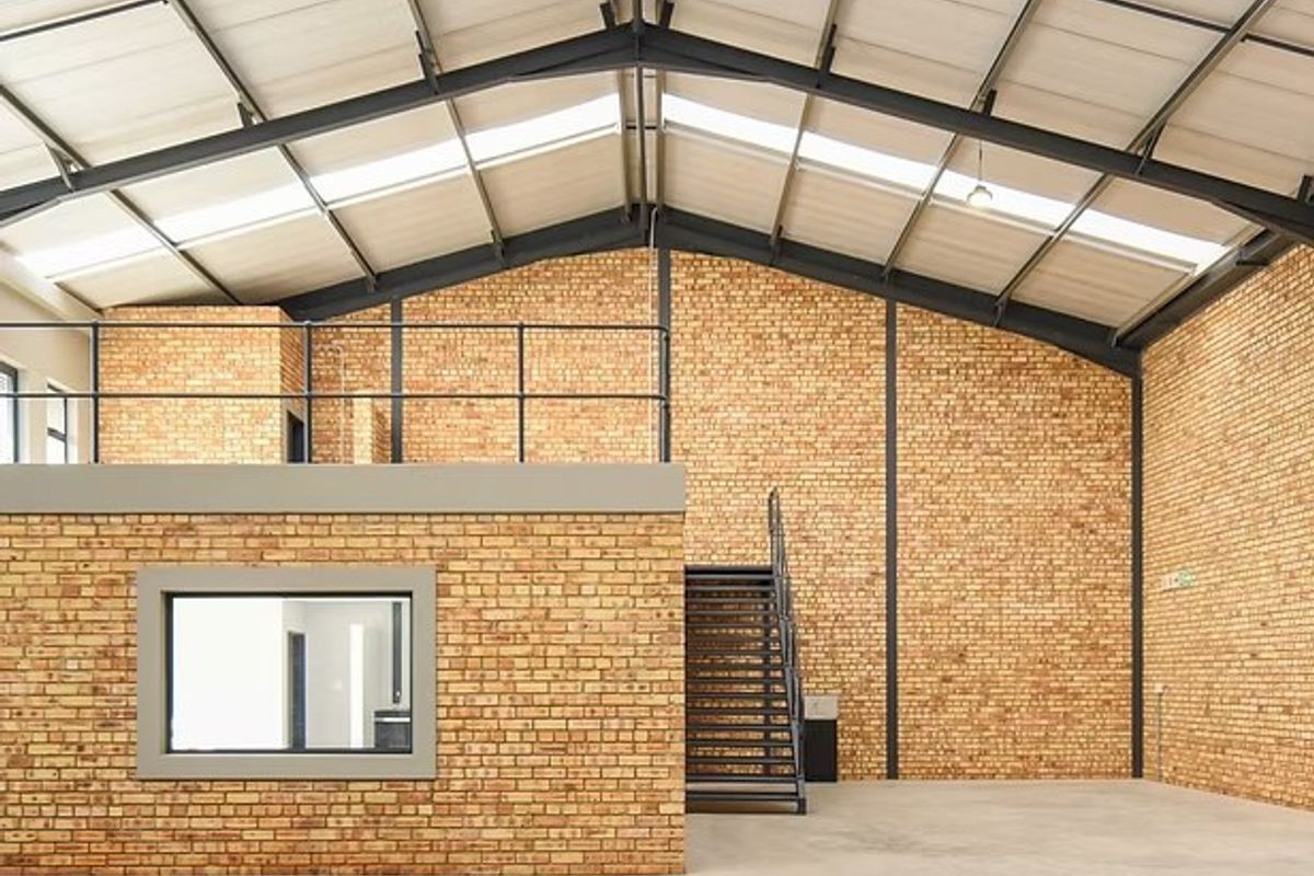 Spacious industrial unit featuring high ceilings with skylights, exposed brickwork, concrete flooring, and a functional mezzanine level accessed by a metal staircase.