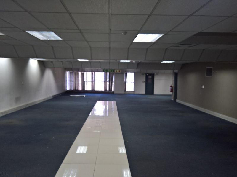 A large, vacant open-plan industrial office space featuring a suspended ceiling, recessed lighting, and a combination of carpeted and tiled flooring.