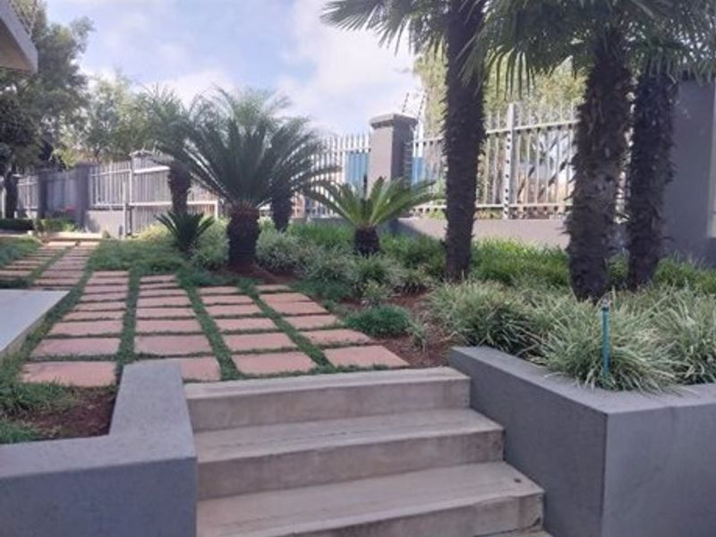 A well-maintained garden area featuring concrete steps, paved pathways, landscaped beds, and secure metal perimeter fencing with integrated electric security wires.