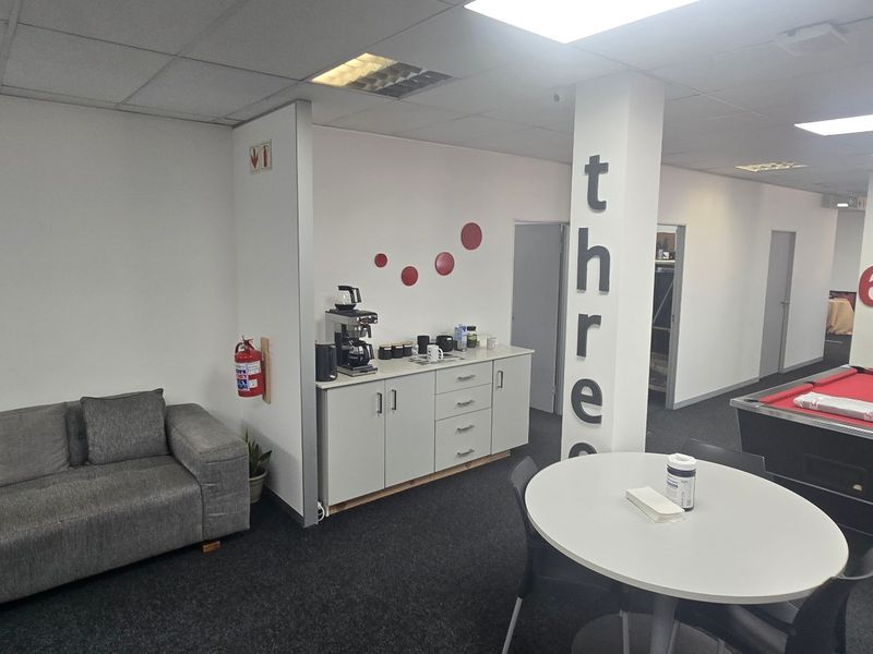 Open-plan commercial office space featuring a kitchenette area, carpeted flooring, and recessed ceiling lighting, suitable for a collaborative work environment.