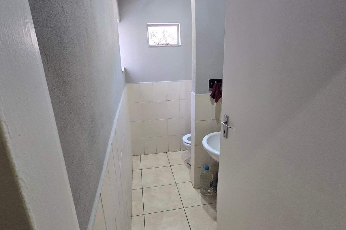 A compact commercial bathroom featuring a pedestal basin, toilet, tiled flooring, and partial wall tiling, requiring cosmetic updates to improve its overall condition.