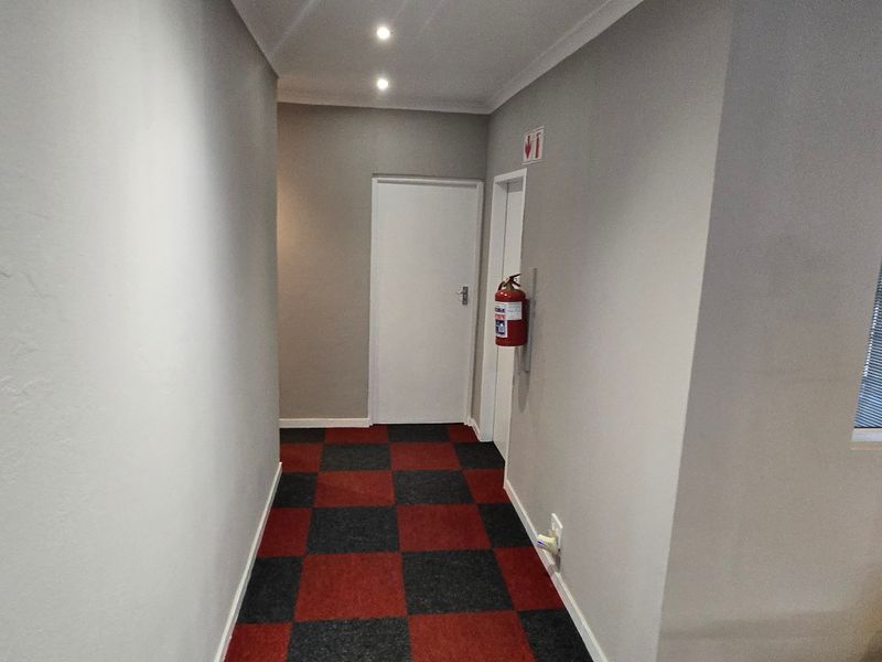 A clean, well-lit commercial hallway featuring checkered carpet tiles, recessed ceiling lighting, and a wall-mounted fire extinguisher.