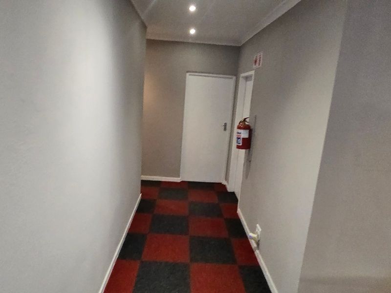 A clean, well-lit commercial hallway featuring checkered carpet tiles, recessed ceiling lighting, and a wall-mounted fire extinguisher.