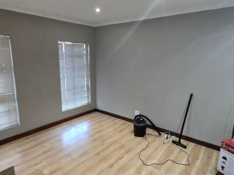 A bright, empty living area featuring light laminate flooring, neutral grey walls, and fitted venetian blinds, offering a clean canvas for office or lounge use.