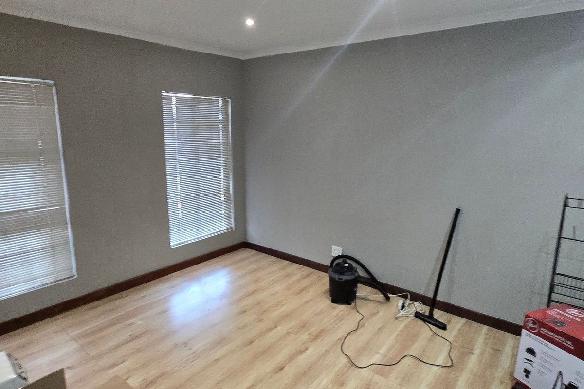A bright, empty living area featuring light laminate flooring, neutral grey walls, and fitted venetian blinds, offering a clean canvas for office or lounge use.