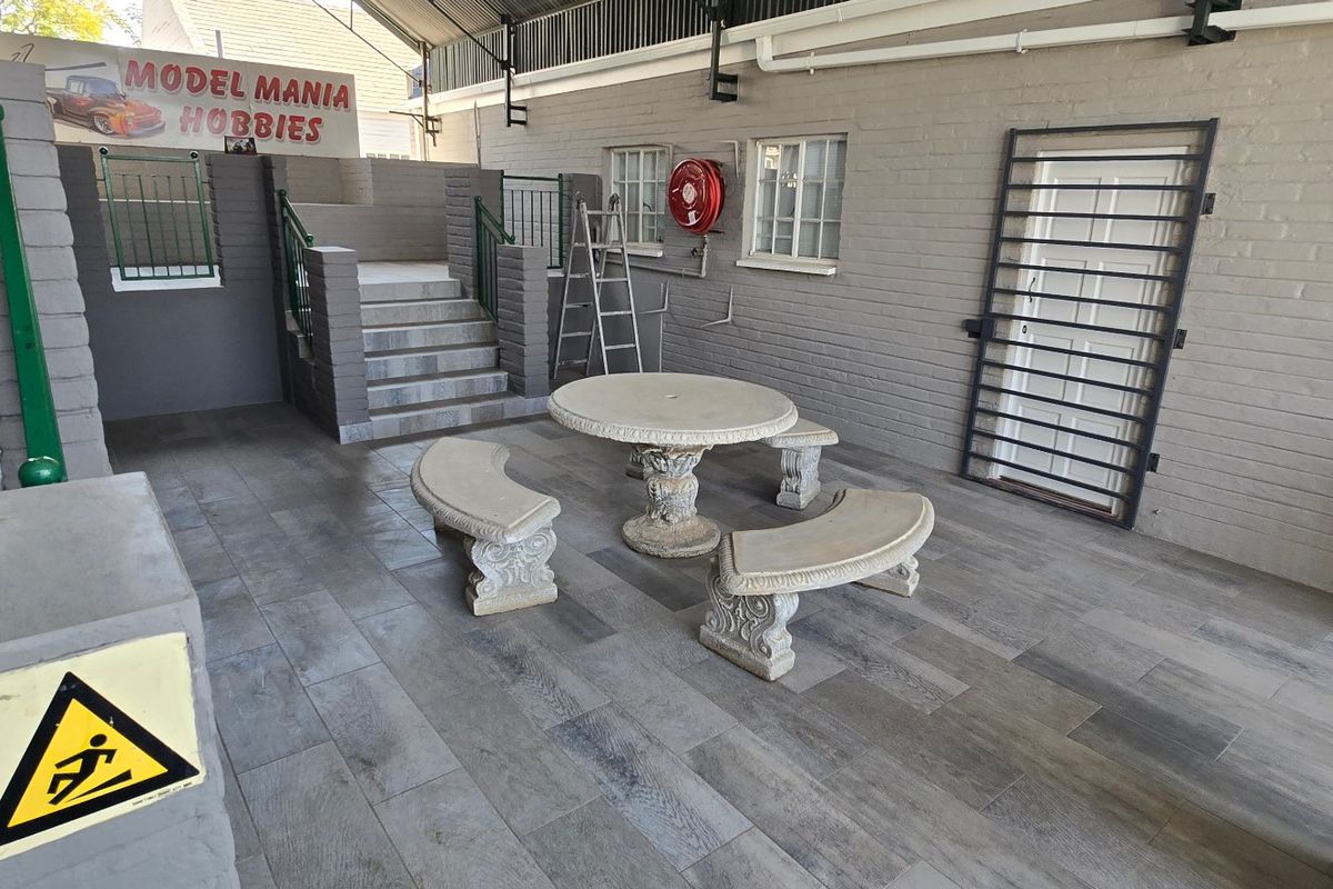 A covered outdoor patio area featuring tiled flooring, brick walls, a security gate, and a staircase leading to an elevated section.