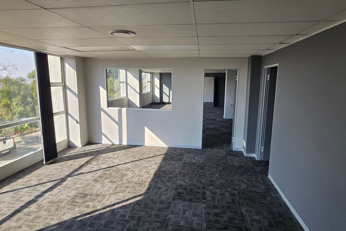 An empty, well-lit commercial office space featuring carpet tiles, a suspended ceiling, and large windows providing natural light and exterior views.