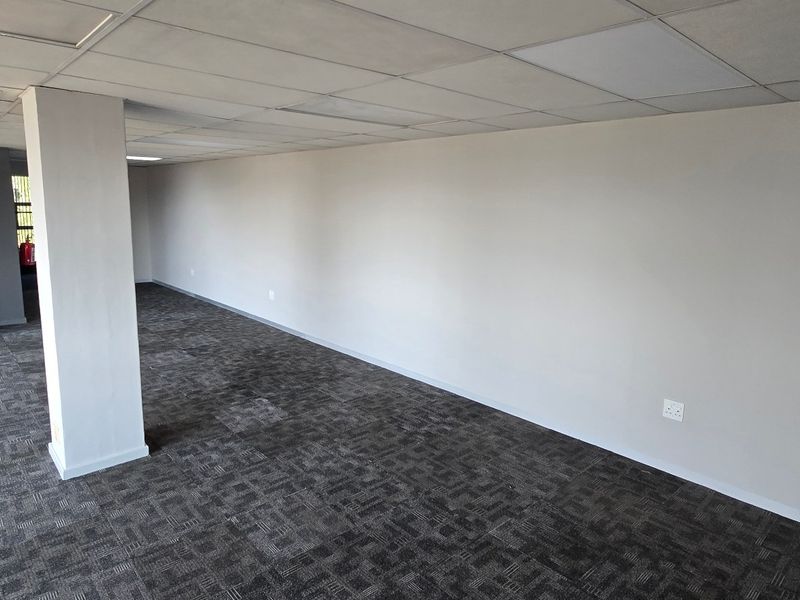 A spacious, empty commercial office area featuring carpet tiles, a suspended ceiling with recessed lighting, and a central air conditioning unit.