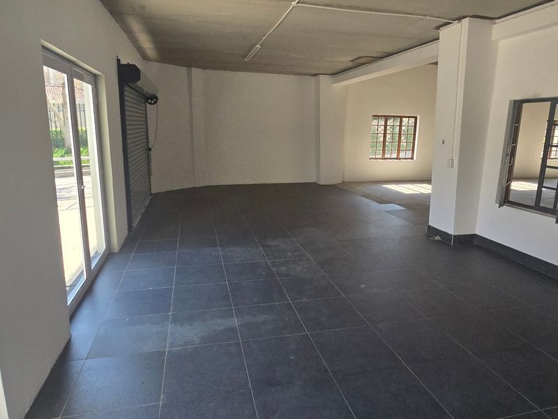 Spacious open-plan industrial unit featuring dark tiled flooring, exposed concrete ceilings, and large metal-framed windows with a secure roller shutter entrance.