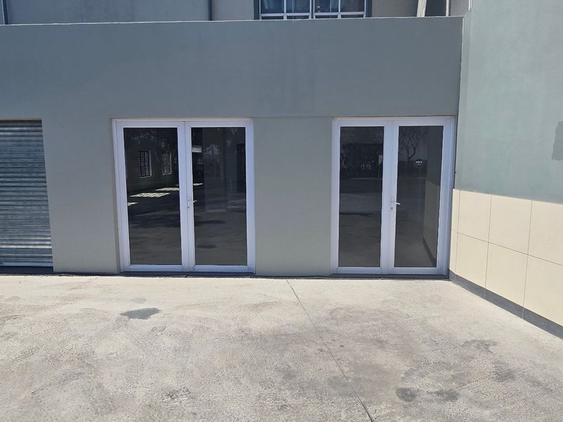 Industrial exterior view featuring a roller shutter door, double glass entry doors, and a concrete driveway area.