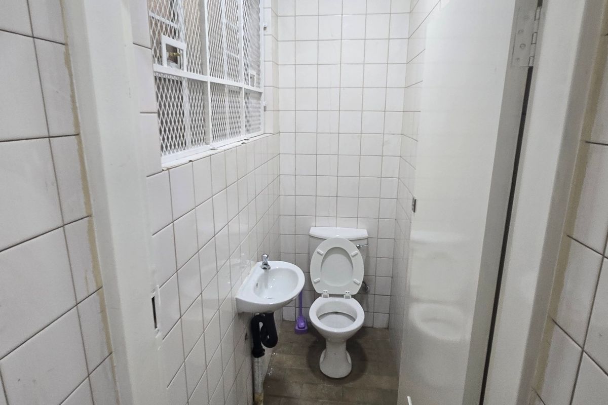 A compact industrial-style bathroom featuring white wall tiles, a wall-mounted basin, and a security grille on the window.