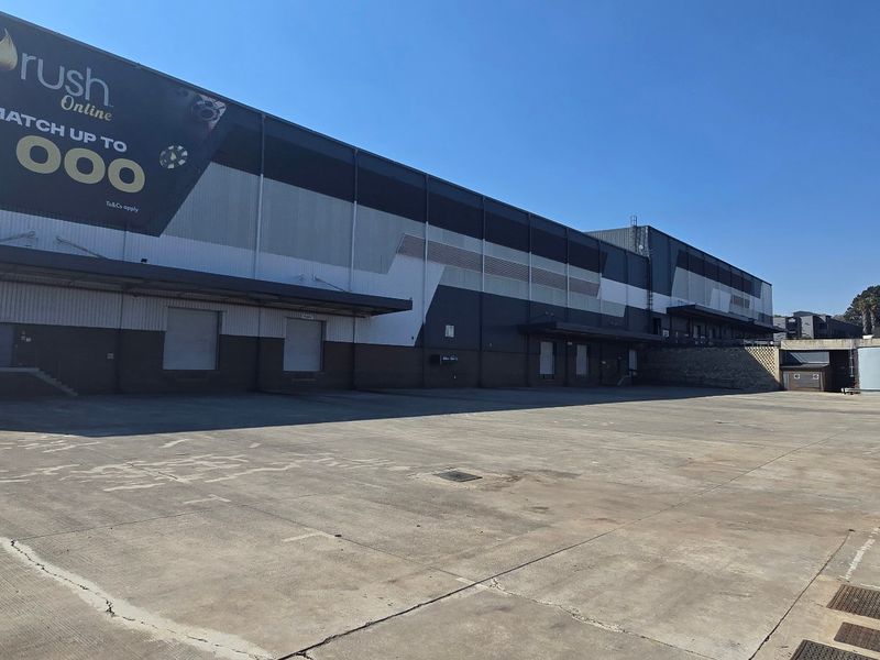 Large industrial warehouse exterior featuring a spacious concrete yard, multiple loading docks, and roller shutter doors, suitable for logistics or manufacturing operations.