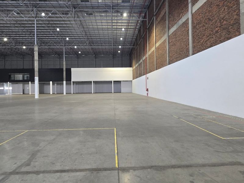A large, open-plan industrial warehouse space featuring high ceilings with exposed steel trusses, concrete flooring, and a combination of brick and painted walls.