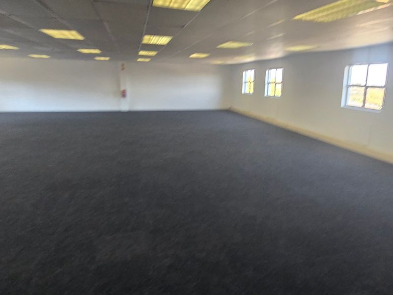 A large, open-plan commercial office space featuring carpeted flooring, a suspended ceiling with recessed lighting, and multiple windows providing natural light.