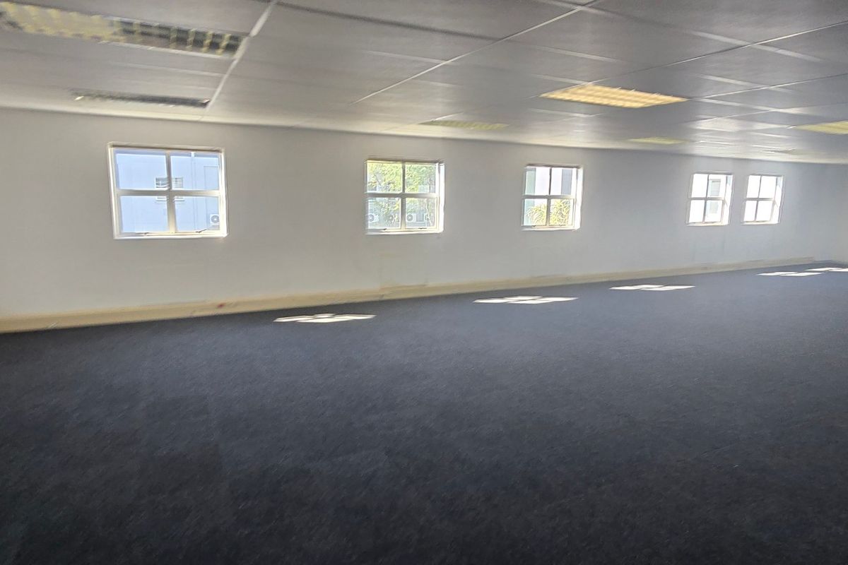 A spacious, open-plan commercial office area featuring neutral walls, dark carpeting, and a suspended ceiling with integrated lighting.
