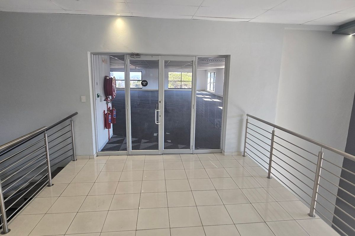 A bright, modern commercial hallway featuring tiled flooring, stainless steel balustrades, and a glass-paneled entrance leading into an open-plan office space.