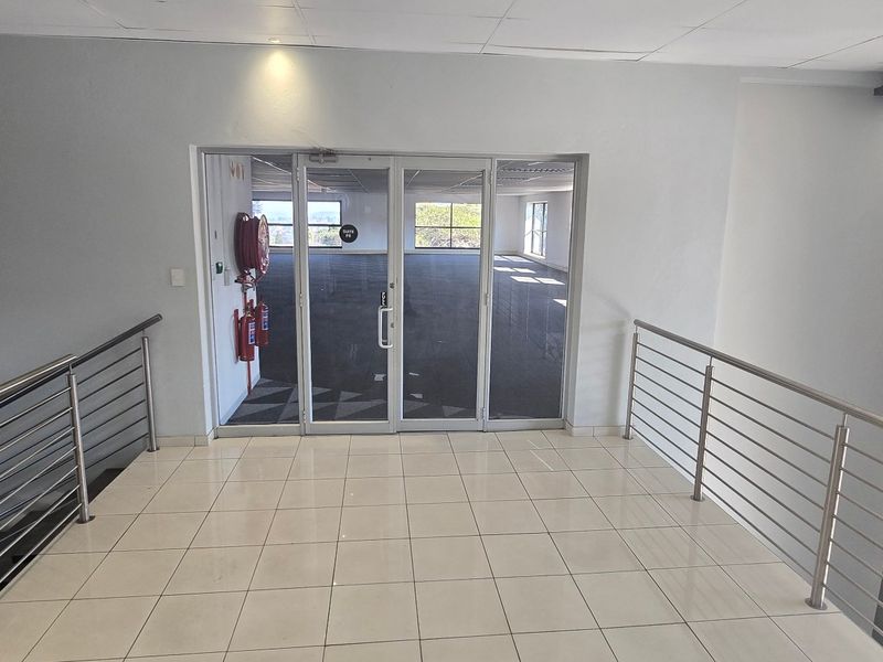 A bright, modern commercial hallway featuring tiled flooring, stainless steel balustrades, and a glass-paneled entrance leading into an open-plan office space.