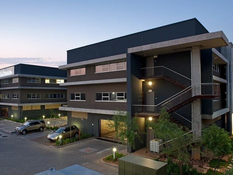 Modern multi-story commercial office building featuring contemporary dark facade, external concrete staircases, and paved parking area with integrated landscaping.