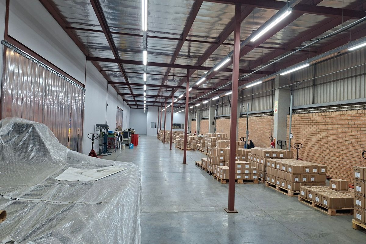 Spacious industrial warehouse area featuring exposed steel roof trusses, concrete flooring, and high-intensity strip lighting, suitable for storage or light manufacturing operations.