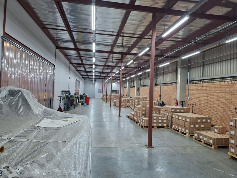 Spacious industrial warehouse area featuring exposed steel roof trusses, concrete flooring, and high-intensity strip lighting, suitable for storage or light manufacturing operations.