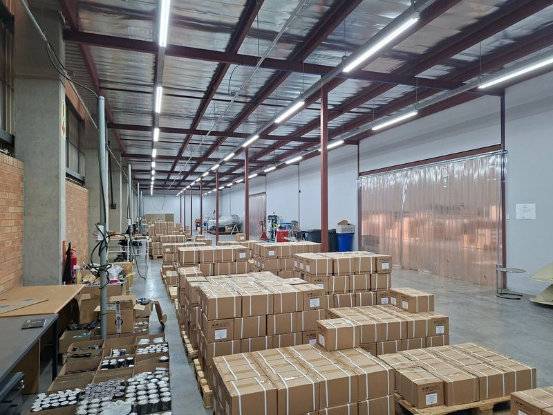 Spacious industrial warehouse area featuring high ceilings with exposed steel beams, fluorescent lighting, concrete flooring, and a practical PVC strip curtain entrance.