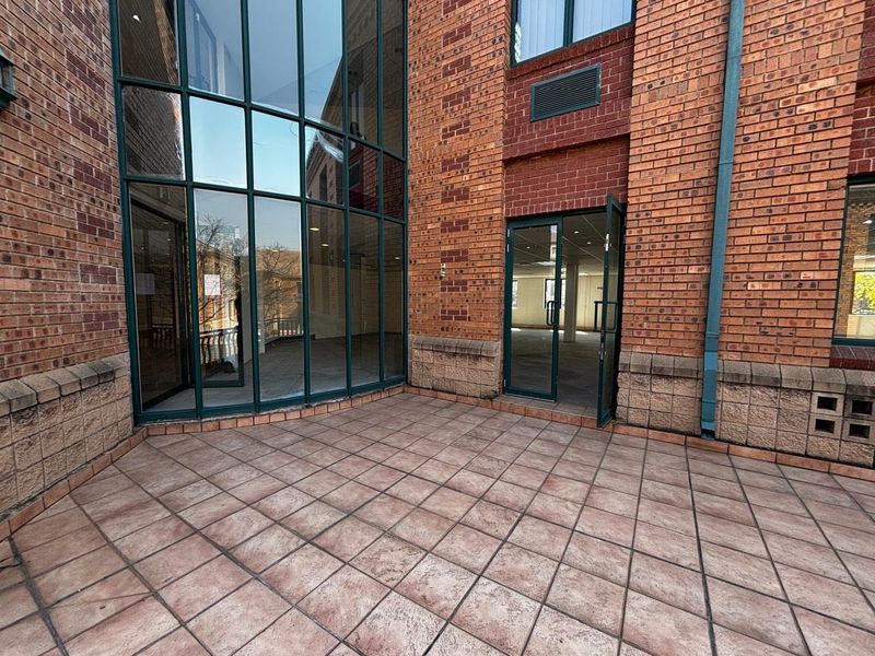 A paved outdoor patio area featuring a full-height glass curtain wall and brick facade, providing direct access to the interior commercial space.