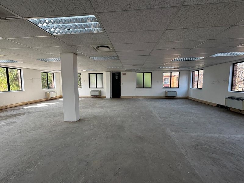 A spacious, open-plan commercial office area featuring a suspended ceiling, recessed lighting, multiple air conditioning units, and unfinished concrete flooring.