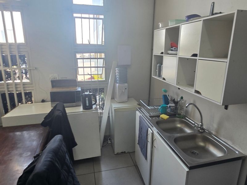 A functional kitchenette featuring a double stainless steel sink, wall-mounted storage cabinets, and security-barred windows, providing basic utility space for an industrial property.