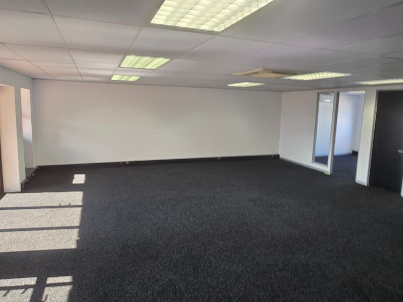 A spacious, empty commercial office area featuring dark carpeting, a suspended ceiling with recessed lighting, and glass-paneled access doors.