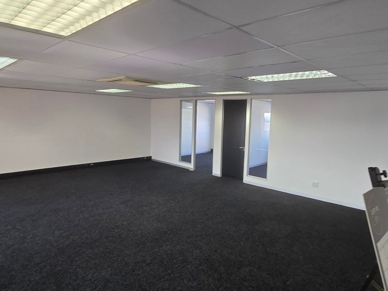 A spacious, empty commercial office area featuring a suspended ceiling with recessed lighting, dark carpeting, and glass-partitioned internal doors.