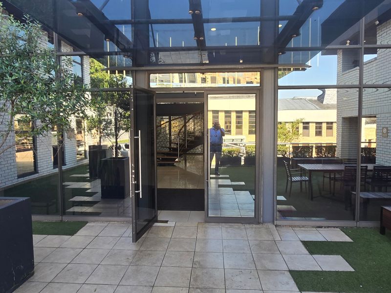 A modern commercial patio area featuring a glass canopy, extensive glass facade, tiled flooring, and integrated artificial turf sections.