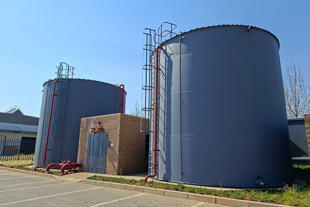 Industrial site featuring two large storage tanks with fixed access ladders and a brick utility building, situated adjacent to a paved parking area.