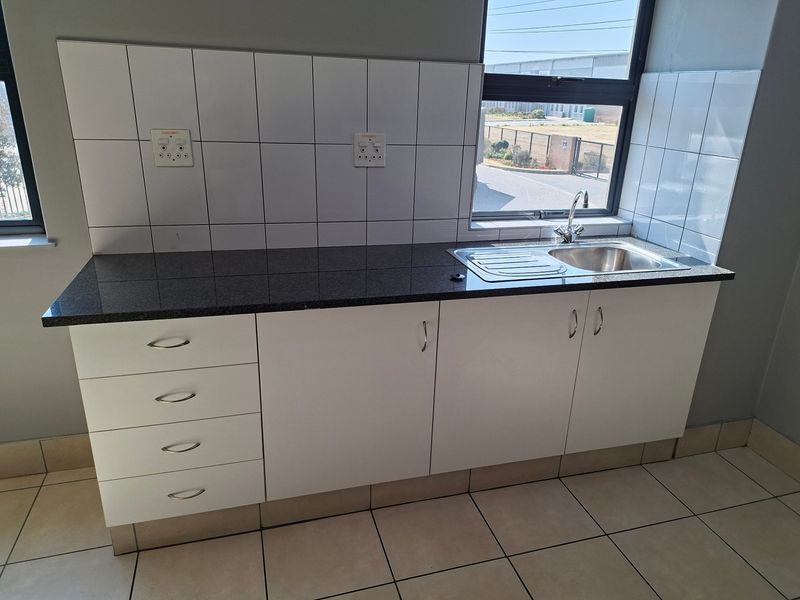 A clean, functional kitchenette featuring granite countertops, white cabinetry, a stainless steel sink, and tiled backsplash in an industrial office setting.