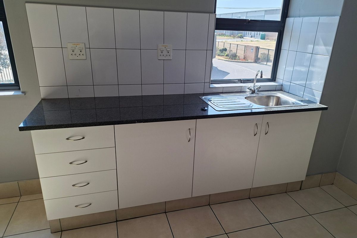 A clean, functional kitchenette featuring granite countertops, white cabinetry, a stainless steel sink, and tiled backsplash in an industrial office setting.