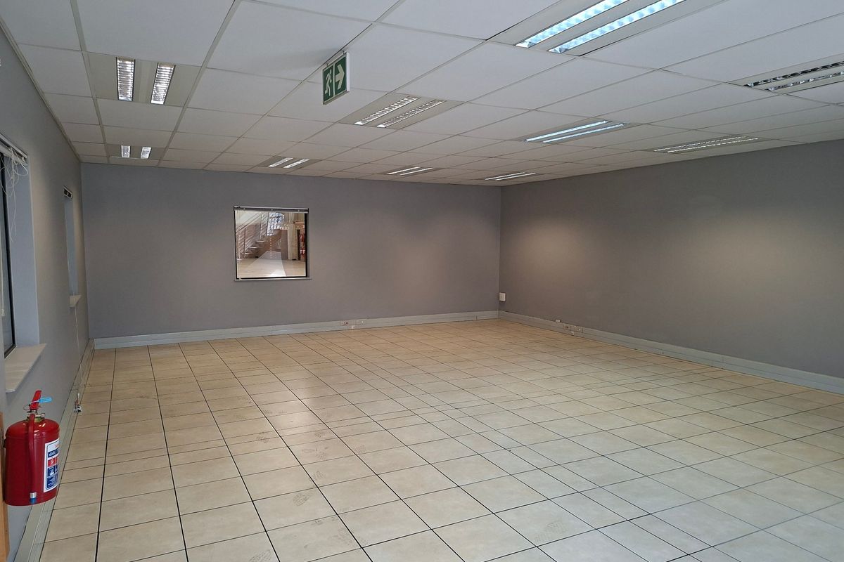 A spacious, empty industrial office area featuring neutral grey walls, ceramic tiled flooring, and a suspended ceiling with recessed lighting.