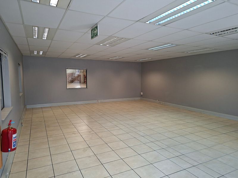 A spacious, empty industrial office area featuring neutral grey walls, ceramic tiled flooring, and a suspended ceiling with recessed lighting.