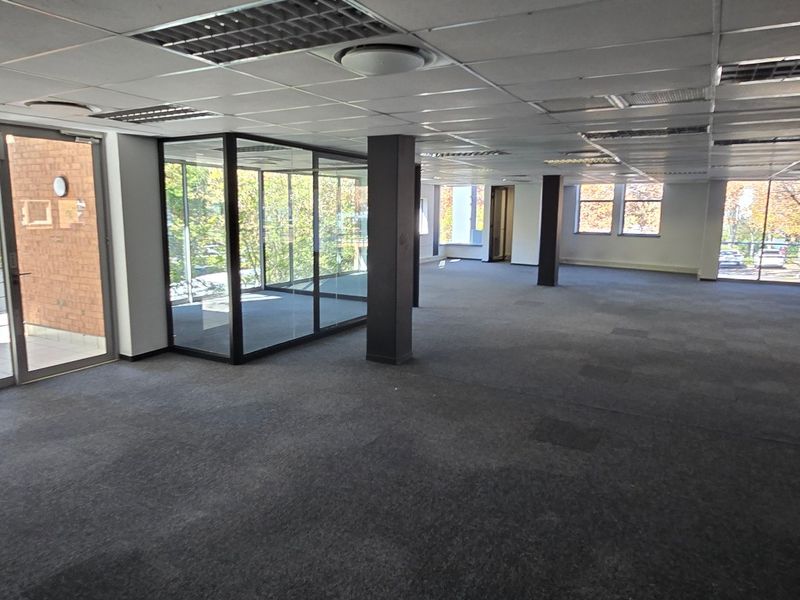Spacious open-plan commercial office area featuring carpeted flooring, suspended ceilings, recessed lighting, and a private glass-enclosed office space with large windows.
