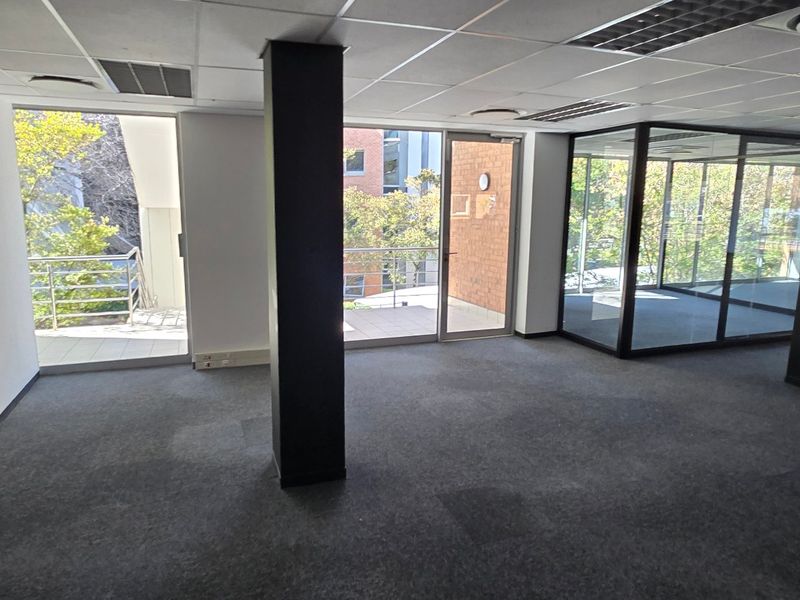 An open-plan commercial office space featuring carpeted flooring, suspended ceilings, structural columns, and glass-partitioned meeting areas with direct access to an outdoor balcony.