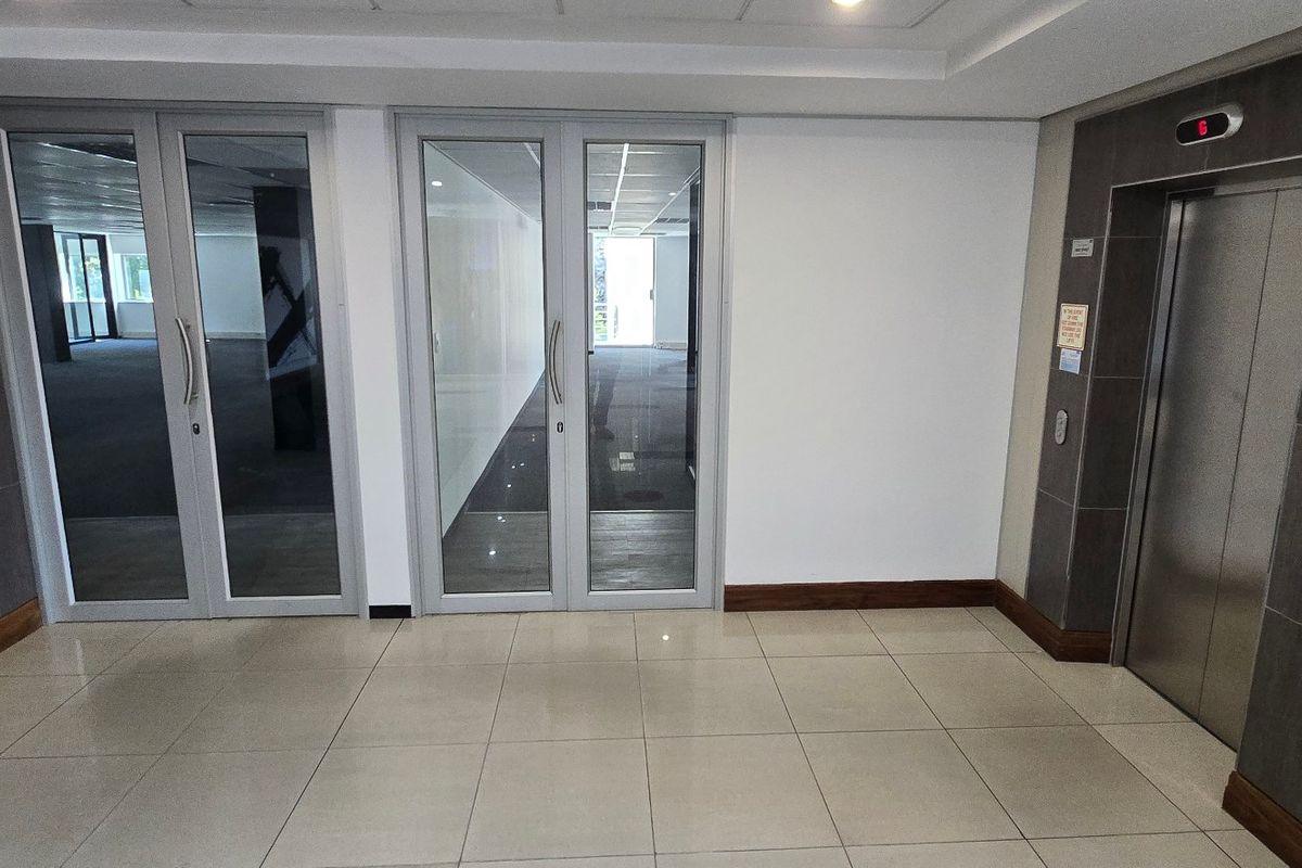 A clean, modern commercial lobby area featuring tiled flooring, an elevator, and glass-panelled double doors leading into the office space.