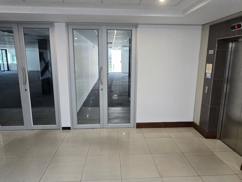 A clean, modern commercial lobby area featuring tiled flooring, an elevator, and glass-panelled double doors leading into the office space.