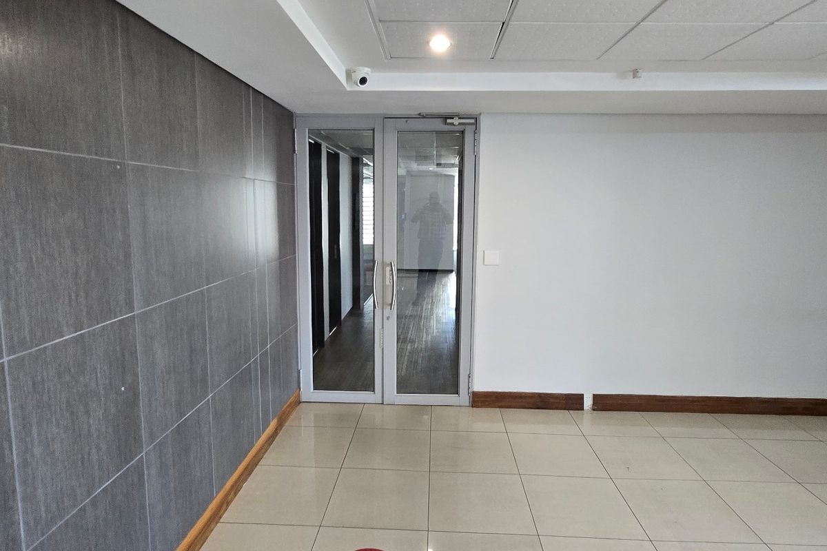 A clean, modern commercial hallway featuring tiled flooring, a suspended ceiling with recessed lighting, and a glass entrance door.