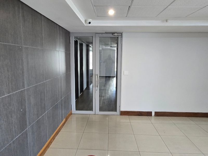 A clean, modern commercial hallway featuring tiled flooring, a suspended ceiling with recessed lighting, and a glass entrance door.