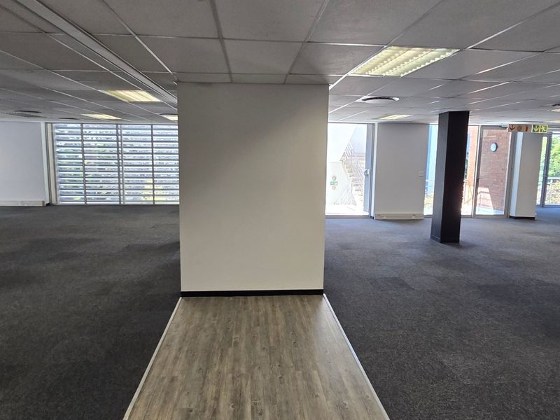 Spacious open-plan commercial office area featuring a mix of carpet and laminate flooring, suspended ceilings, and large windows providing ample natural light.