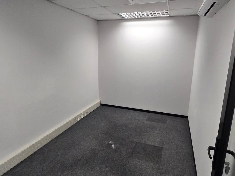 A vacant, compact office space featuring carpeted flooring, a suspended ceiling with recessed lighting, and a wall-mounted air conditioning unit.
