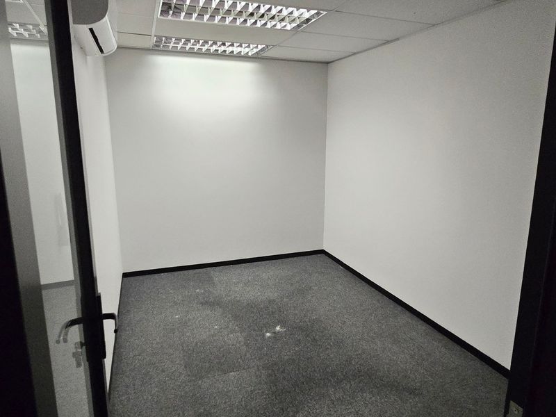 A vacant, neutral-toned office space featuring carpeted flooring, a suspended ceiling with recessed lighting, and a wall-mounted air conditioning unit.