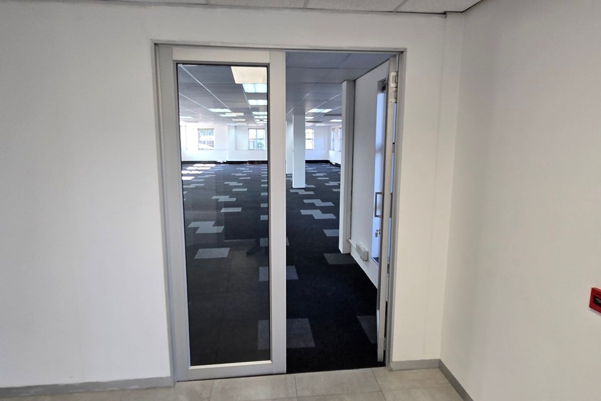An open-plan commercial office space featuring carpet tiles, suspended ceilings with recessed lighting, and a modern aluminium-framed glass entrance door.