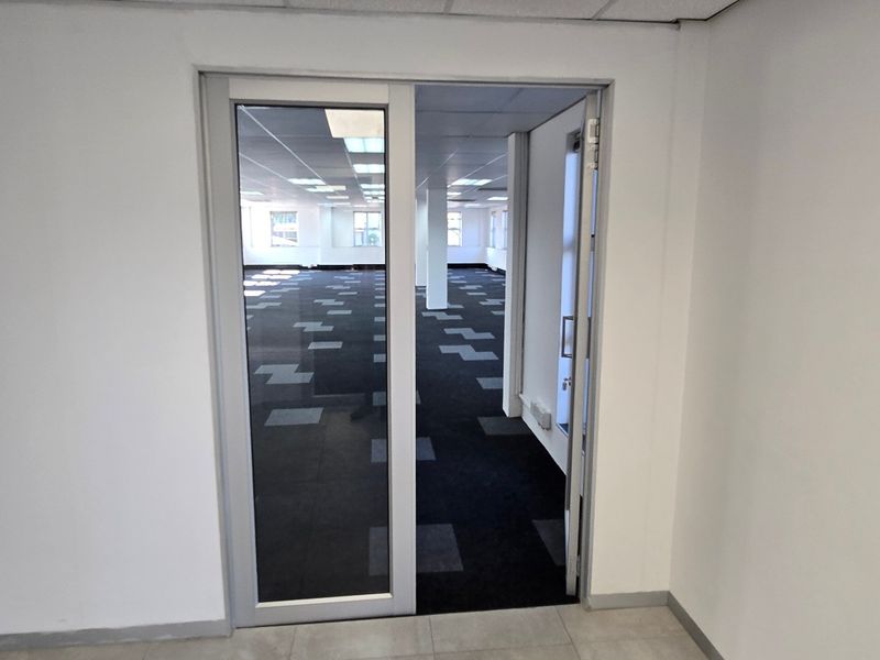 An open-plan commercial office space featuring carpet tiles, suspended ceilings with recessed lighting, and a modern aluminium-framed glass entrance door.