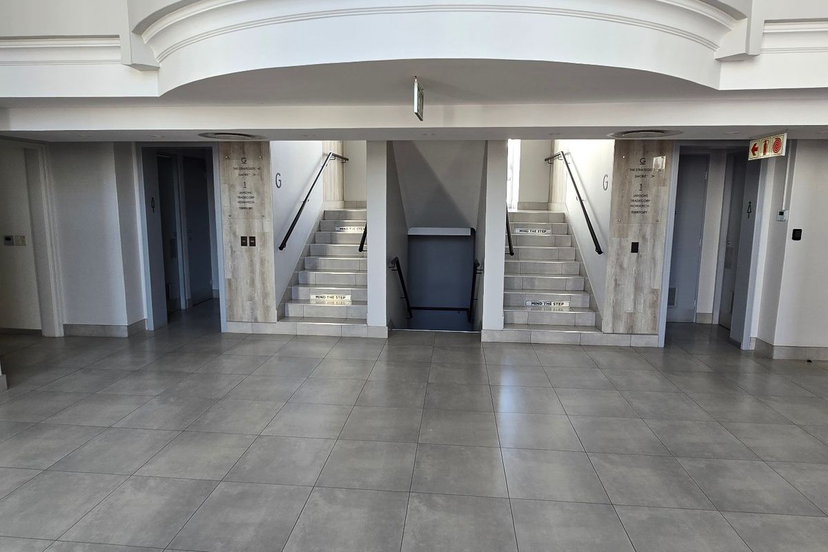 A clean, modern commercial building lobby featuring tiled flooring, dual staircases with handrails, and clear directional signage.