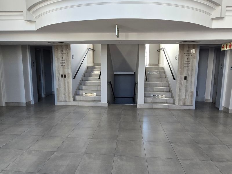 A clean, modern commercial building lobby featuring tiled flooring, dual staircases with handrails, and clear directional signage.